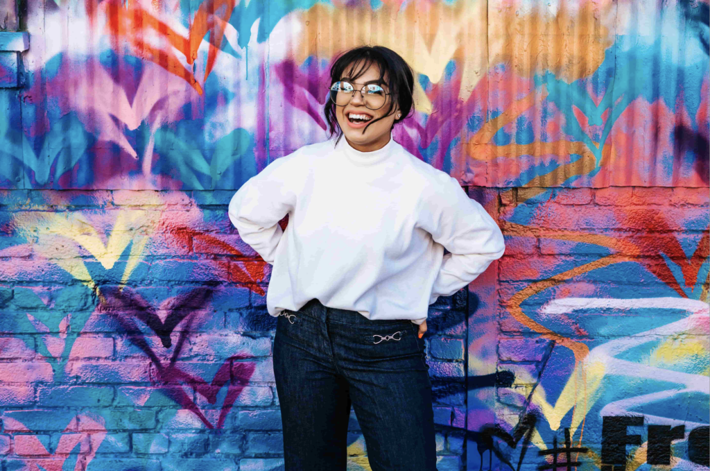 positive body affirmation post: woman wearing glasses smiling in front of street art